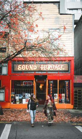 Two people walking past a red storefront with autumn leaves on the ground.