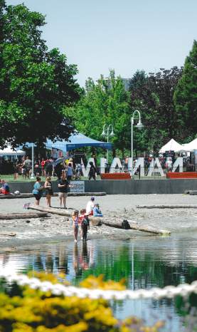 People gather along a waterfront park in Nanaimo (Maffeo Sutton Park), with children wading in the water and market tents and the ‘Nanaimo’ sign in the background.