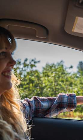 Smiling Woman in Plaid Shirt Driving Past Orchard with Hand Out Car Window