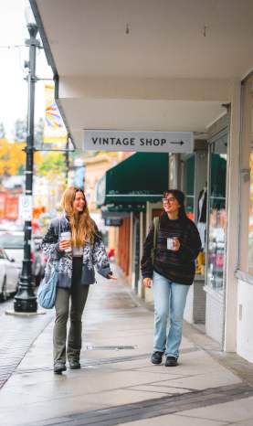 Two people stroll past shops on a rainy autumn day in downtown Nanaimo, holding coffee as colourful fall trees line the street.