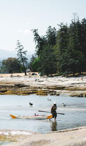One person paddles a kayak while another walks beside a kayak in shallow water along a rocky shoreline.