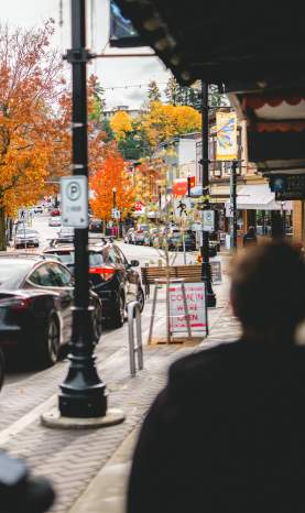 People walk along a downtown Nanaimo street with shops, parked cars, and vibrant autumn trees lining the road.