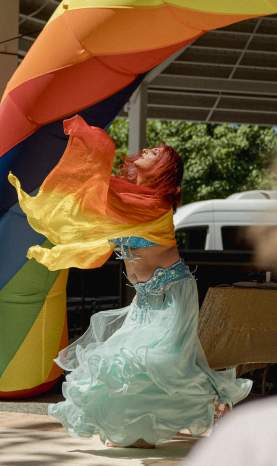 Performer in flowing costume dancing beneath a rainbow arch on a stage at a Pride event, with audience in the foreground.