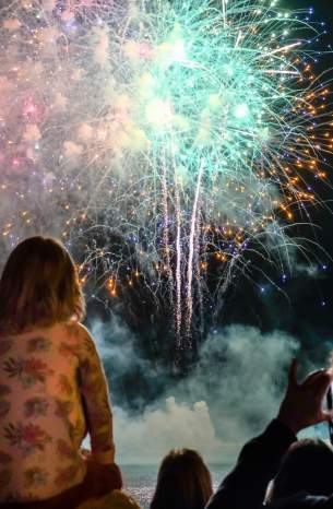 A child sits on their parent's shoulders watching a fireworks show