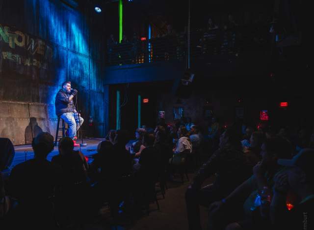 A man sitting on a stool onstage with a mic during a comedy set at Vulcan Gas Company.