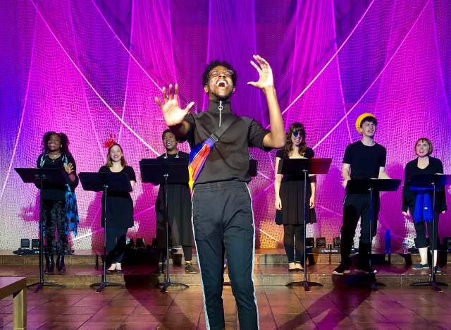 photo of a performance at Trinity Street Playhouse. A man wearing black athletic clothing stands in the center, singing, and a chorus stands behind him. Each chorus member is singing and stands behind a black music stand