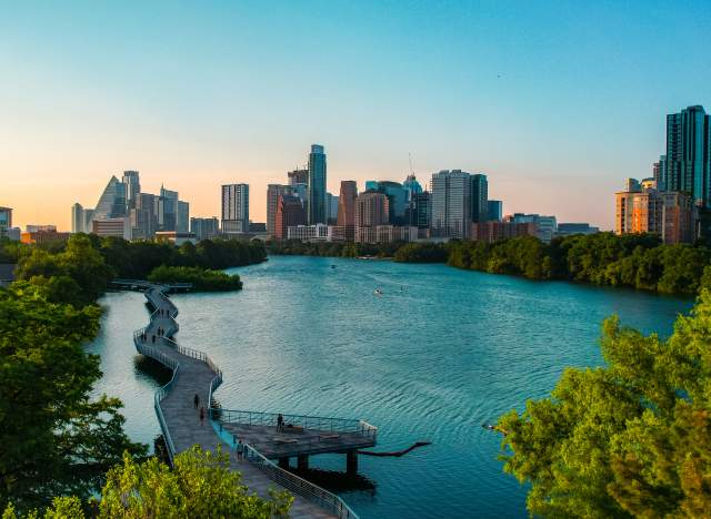 People walking along suspended board walk that follows the shape of the Colorado River, leading to Downtown Austin skyline.