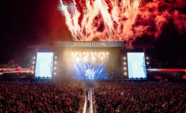 Photograph showing the culmination of a headline set at Victorious Festival, with red fireworks going off behind the stage and thousands of people gathered in front