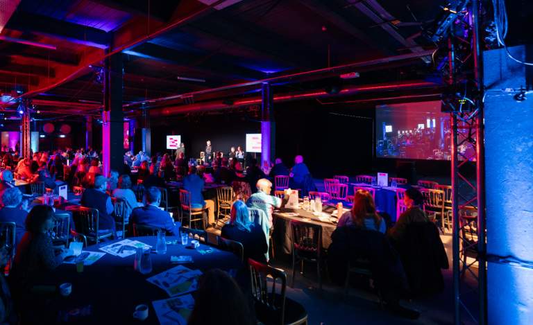 Audience watching a conference stage which is bathed in red and purple light.