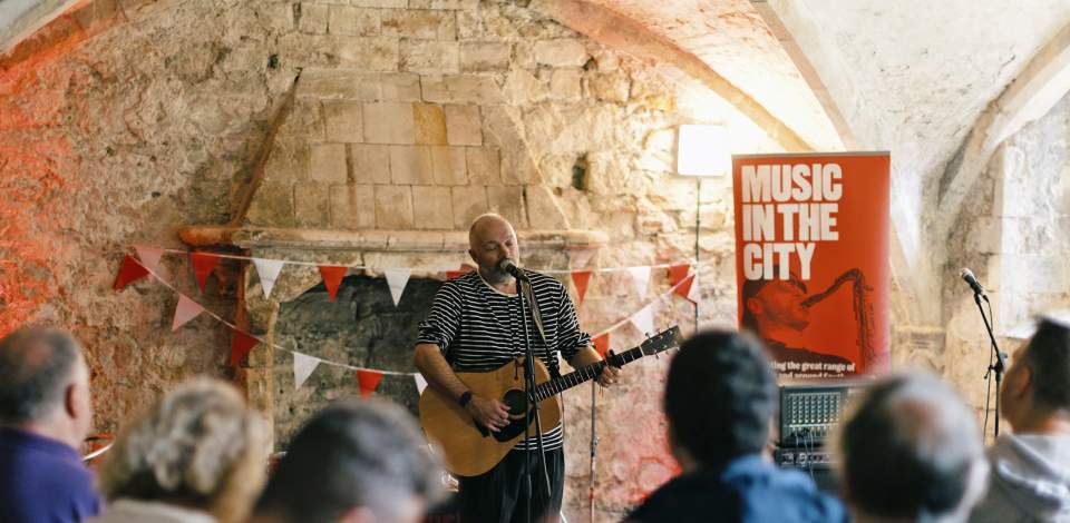 Man singing into a mic whilst playing an acoustic guitar in front of a crowd, inside a Southampton vault for Music In The City 2025