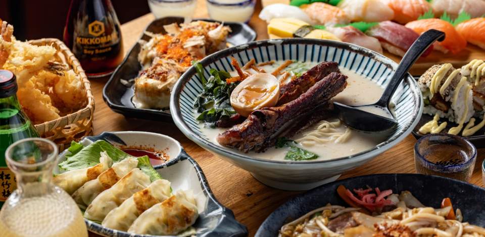 Spread of Japanese cuisine on wooden table including big bowl of ramen, dumplings, and sushi