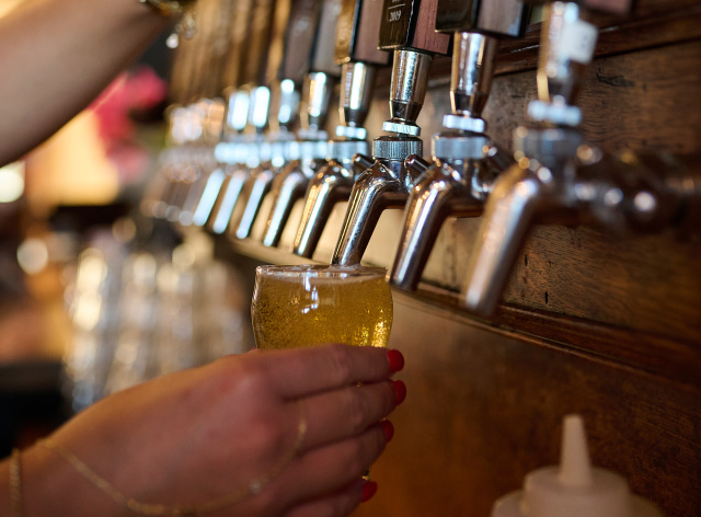 Close-up of a bartender pouring a golden beer from a row of draft taps labeled with Milwaukee Brewing Company beers into a small tasting glass.