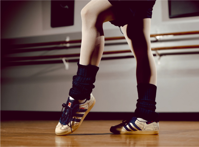 Close-up of a dancer’s legs and feet in sneakers and leg warmers, poised on a wooden studio floor near a ballet barre.
