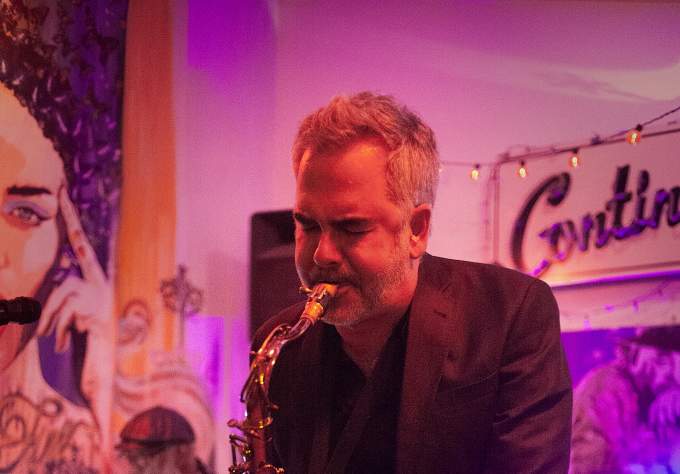 Musician Elias Haslanger wearing a black suit and playing the tenor saxophone while leaning toward the camera
