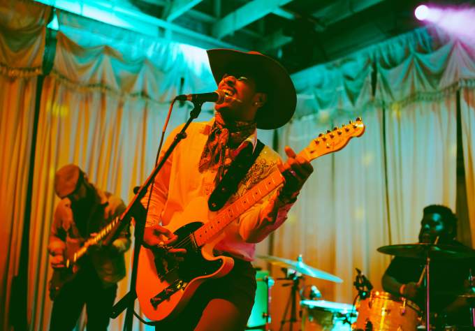 Musician in a cowboy hat singing into a mic while playing guitar with colorful lights surrounding him.