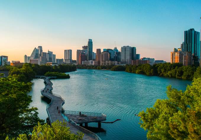 People walking along suspended board walk that follows the shape of the Colorado River, leading to Downtown Austin skyline.