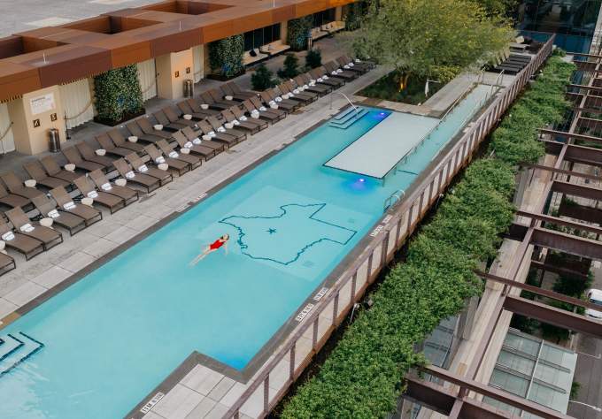 Aerial photo of the JW Marriott rooftop pool with a woman in a red bathing suit swimming on her back in the center of the pool. The floor of the pool includes an outline of the state of Texas with a star on the location of Austin.
