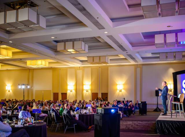 Will Guiderra, a white man in a suit, standing on stage talking to a room full of people sitting at round tables