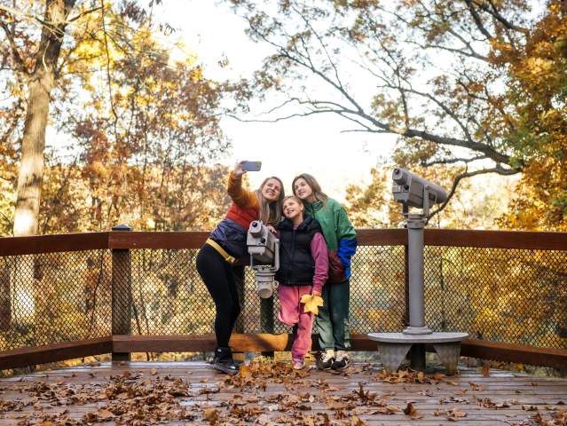 Family smiles and poses for a selfie outdoors during a fall day.