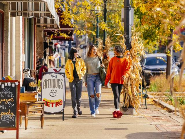 Three women shopping in Depot Town, walking along storefronts on a sidewalk during the fall season. Yellow and orange leaves in the background.