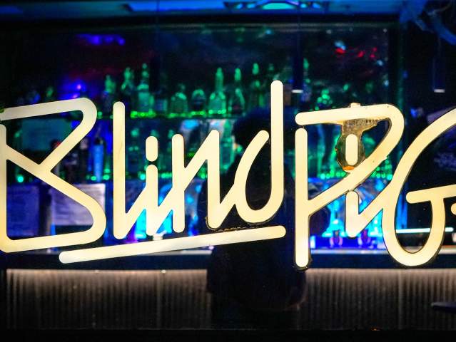 Window cling that reads "Blind Pig". Inside of the glass, a person stands at a dimly-lit bar.