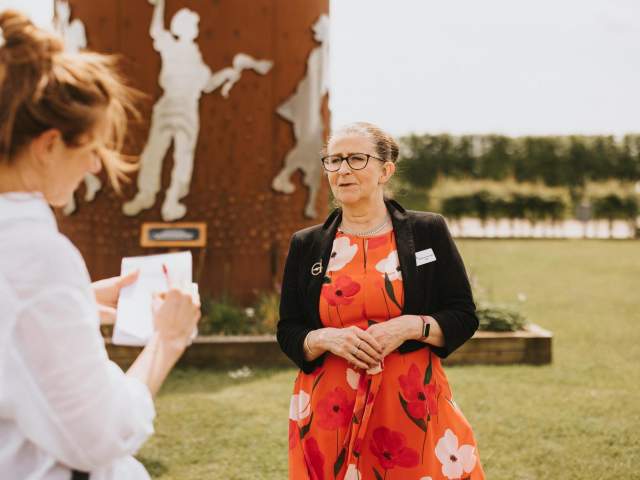 Woman in red dress looking at woman in white shirt in front of a steel sculpture outdoors.