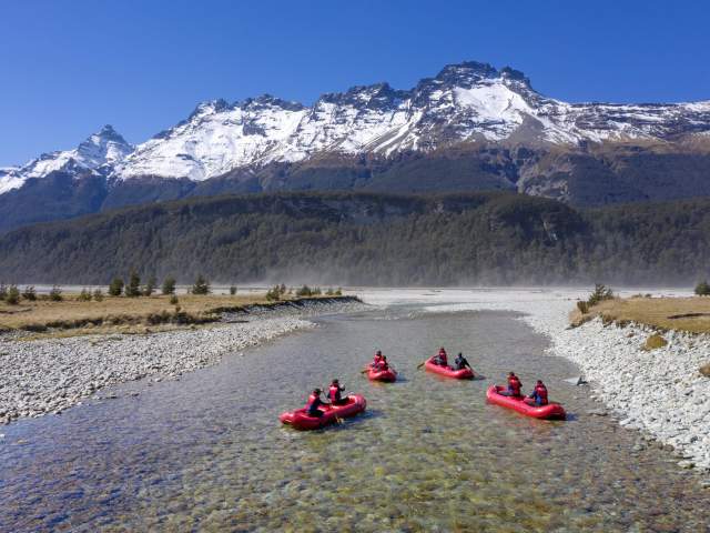 Group of people on blow up kayaks floating down river stream with snow-capped mountains in background