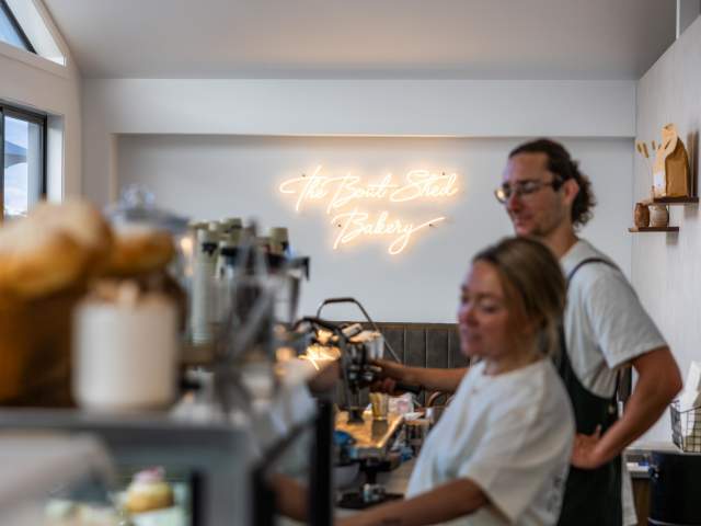Two barisstas making coffee, with a neon sign reading The Boatshed Bakery in the background