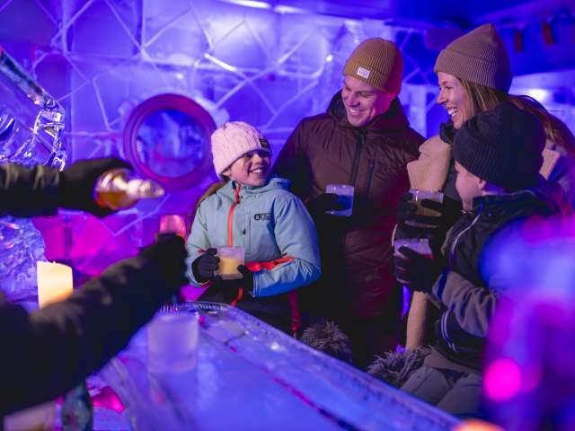A family of four enjoying drinks in an ice bar, surrounded by illuminated ice sculptures and glowing purple and blue lighting. They are dressed warmly in coats, hats, and gloves, smiling and sharing a joyful moment.