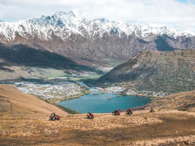 A scenic view of Queenstown, New Zealand, captured from a high vantage point. The image shows snow-capped mountains in the background, a town nestled by a serene lake in the valley, and a group of quad bikes traversing a grassy hill in the foreground.