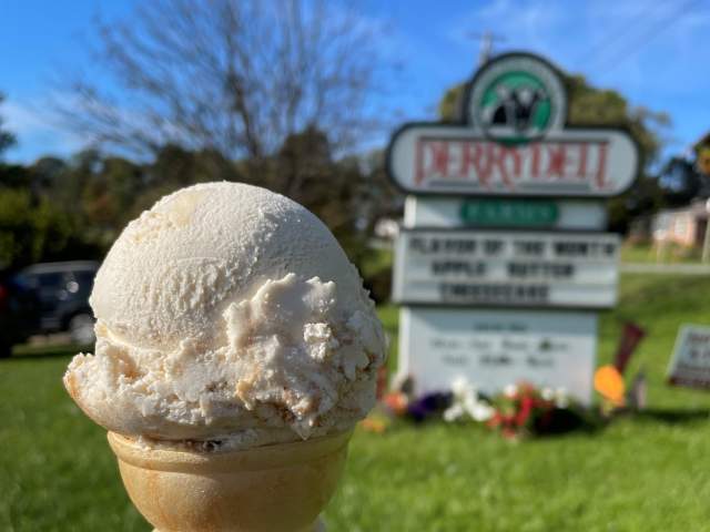 A hand holding up an ice cream cone with one scoop of vanilla ice cream in front of a sign that reads Perrydell with ice cream specials listed underneath