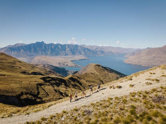 A group of hikers climbing the top of a mountain with views of Lake Whakatipu and Remarkable Mountains in the background