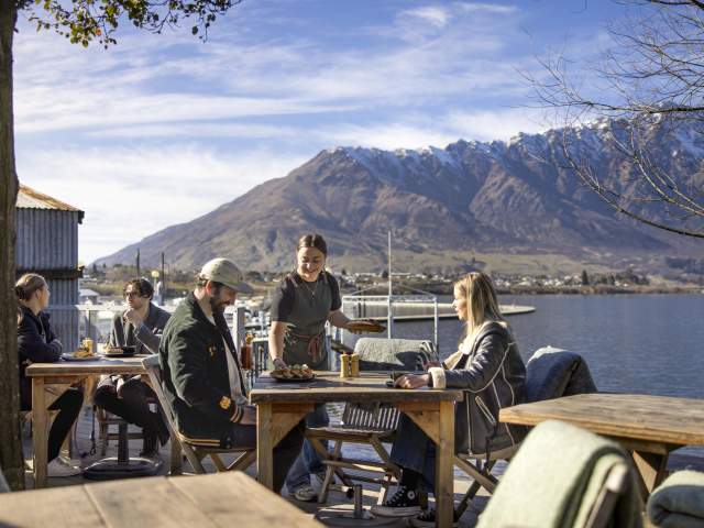 People dining outside next to the lake with the Remarkables mountain range in the background at The Boat Shed