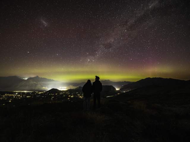 People standing on a mountain top with a starry night sky and aroura before them