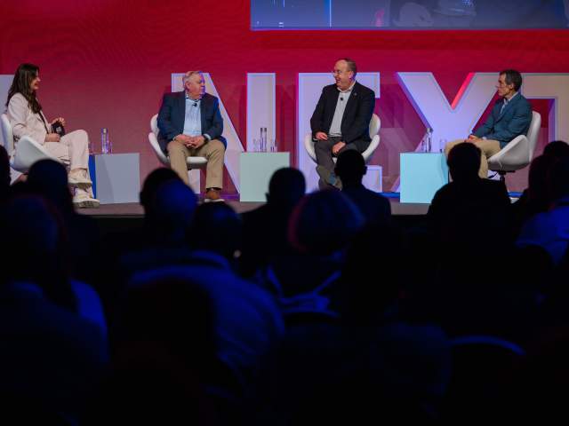 A woman sitting on a stage with three men to her right, speaking on a panel in front of a dark room of attendees.