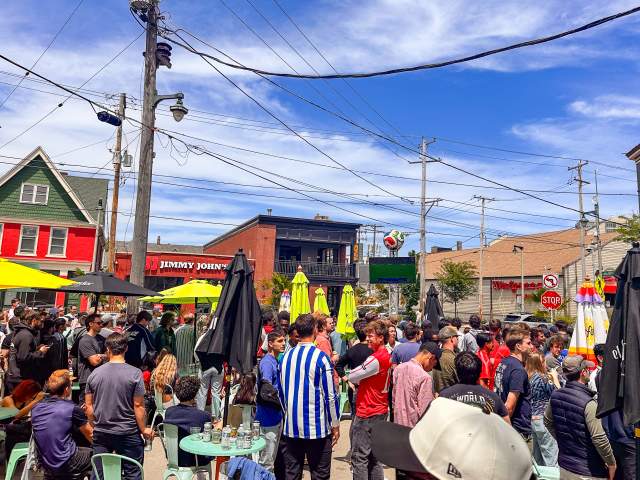 Soccer fans pack the patio and surrounding area outside Nomad World Pub on Brady Street in Milwaukee, watching a match on a large outdoor screen during a sunny afternoon.