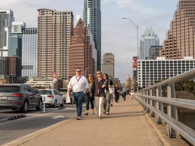 A group of people wearing SXSW badges walk across the Congress Avenue Bridge with the Austin skyline in the background.