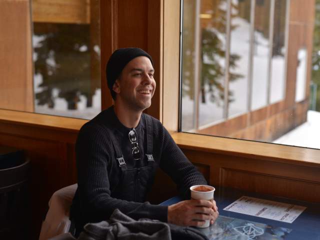 A man in snow bibs sits by a window with snow outside drinking a hot cocoa