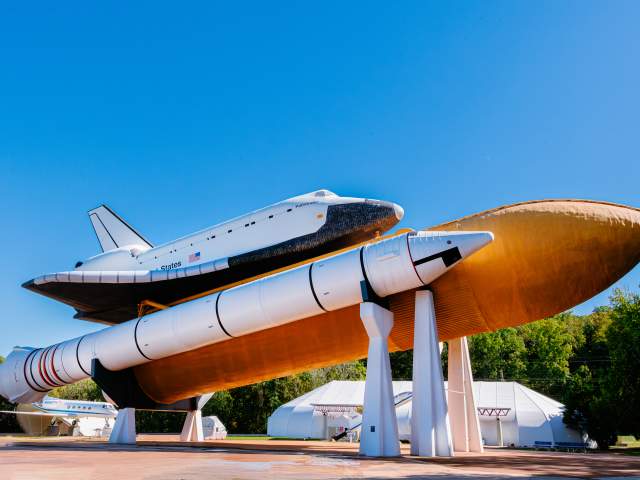 The Space Shuttle Pathfinder is displayed atop a full-scale external tank and solid rocket boosters at the U.S. Space & Rocket Center in Huntsville. The shuttle and rocket stack stand against a clear blue sky, showcasing the scale and engineering of NASA’s Space Shuttle program.