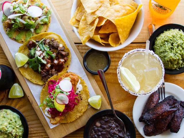 spread featuring assorted street tacos, tortilla chips, guacamole, black beans, cilantro rice, roasted chicken, margaritas, salsa, and fresh lime wedges arranged on a wooden table.