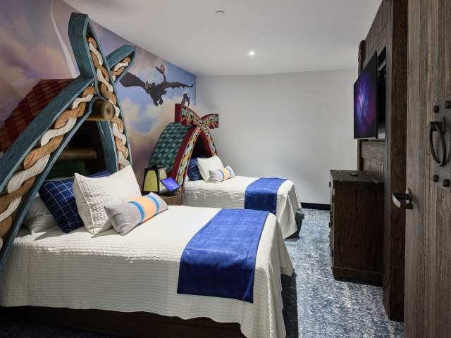Two beds with carved wooden headboards, a dragon wall mural, and rustic wooden elements in the How to Train Your Dragon Kids Suite at Universal Helios Grand Hotel, a Loews Hotel