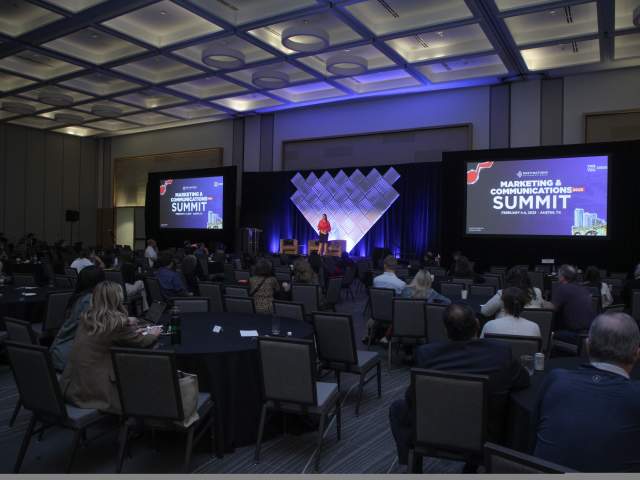 Woman standing on stage at DI Marketing & Communications Summit 2025, giving a welcome message to attendees in a ballroom.