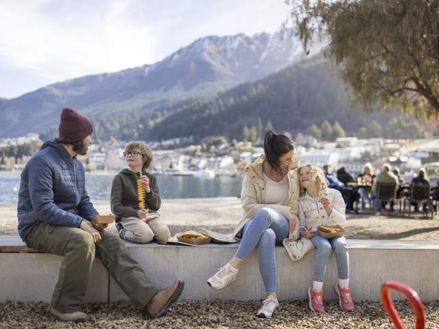Family enjoying fish and chips by Queenstown waterfront with mountains in the background