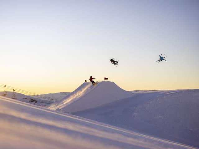 Snowboarders and a skier perform aerial tricks off a jump at Cardrona Alpine Resort at sunrise.