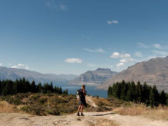 A girl standing at the top of Queenstown Hill after hiking up. She is looking out to the view of the town, Lake Whakatipu and mountain ranges.
