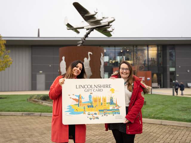 Two people holding a large Lincolnshire Gift Card in front of the International Bomber Command Centre