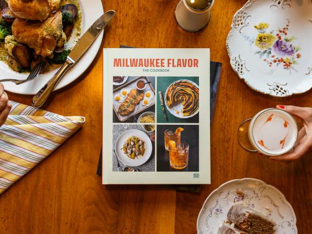 Milwaukee Flavor: The Cookbook rests on a wooden dining table surrounded by a meal. A hand holds a fork over a plate of chicken, vegetables, and potatoes on the left, while another hand holds a latte with heart-shaped foam art on the right. Around the book are a striped napkin, a floral plate, and a dessert plate with slices of cake.