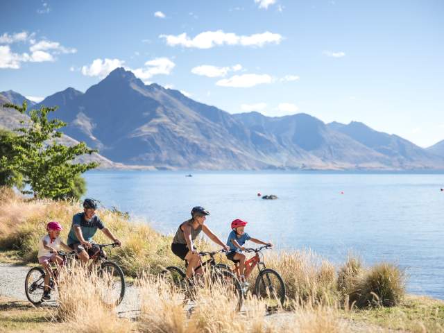 A Family biking around the Queenstown Gardens, with Lake Whakatipu and Cecil Peak in the background