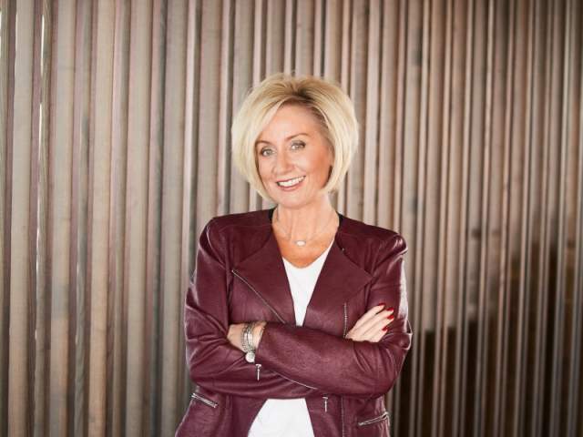 A woman with short blond hair stands smiling with her arms crossed in front of a wall of vertical wooden slats. She wears a burgundy leather jacket over a white top and black pants.