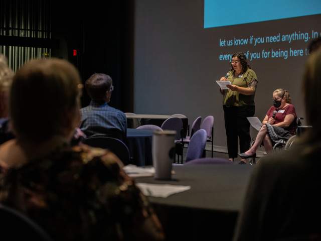 two presenters address a crowd, one standing and one utilizing a wheelchair